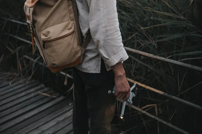 A man wearing a backpack stands on a wooden walkway holding a drone, surrounded by tall grass